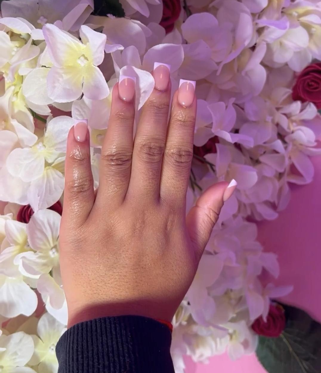 Een hand met roze French nagels tegen een muur van witte en roze bloemen